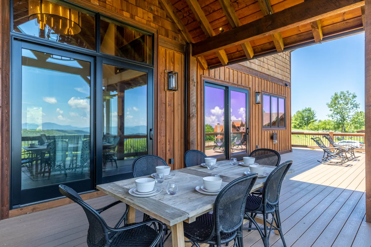 An inviting outdoor dining area featuring a sturdy wooden table surrounded by black wicker chairs, set on a covered deck. Large windows provide a seamless transition between indoors and out, while the background reveals lush mountain views, creating a sense of tranquility and connection to nature.