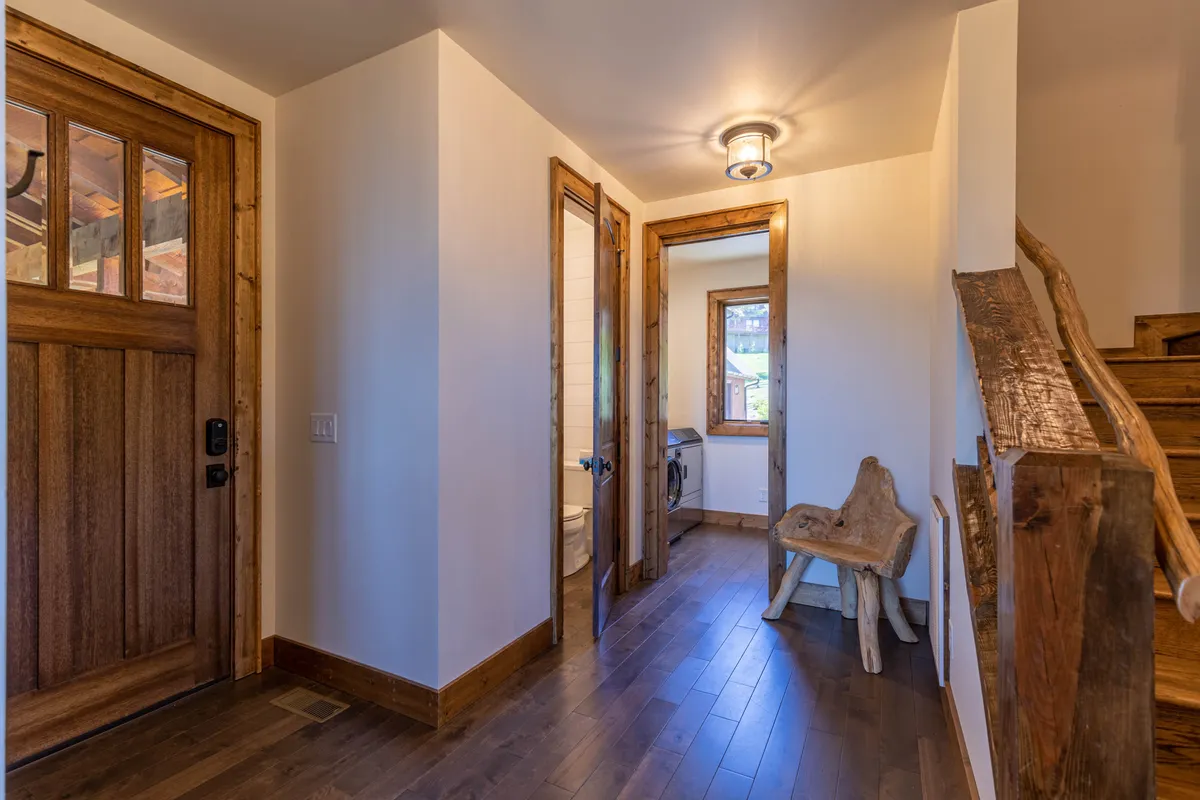 A rustic yet refined entryway featuring a wooden front door, rich wooden staircase, and a natural wooden bench. Framed windows invite natural light, adding warmth and a welcoming ambiance to the space.