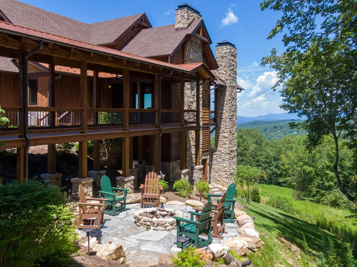 The image showcases the exterior of Breezeway Lodge, prominently featuring a spacious stone patio with Adirondack chairs encircling a glowing fire pit. Wooden pillars frame the patio, while expansive mountain vistas stretch into the distance. The scene is set at sunset, with warm, inviting light and a sense of tranquility. The setting suggests opportunities for stargazing, sharing stories by the fire, or simply soaking in the breathtaking views.
