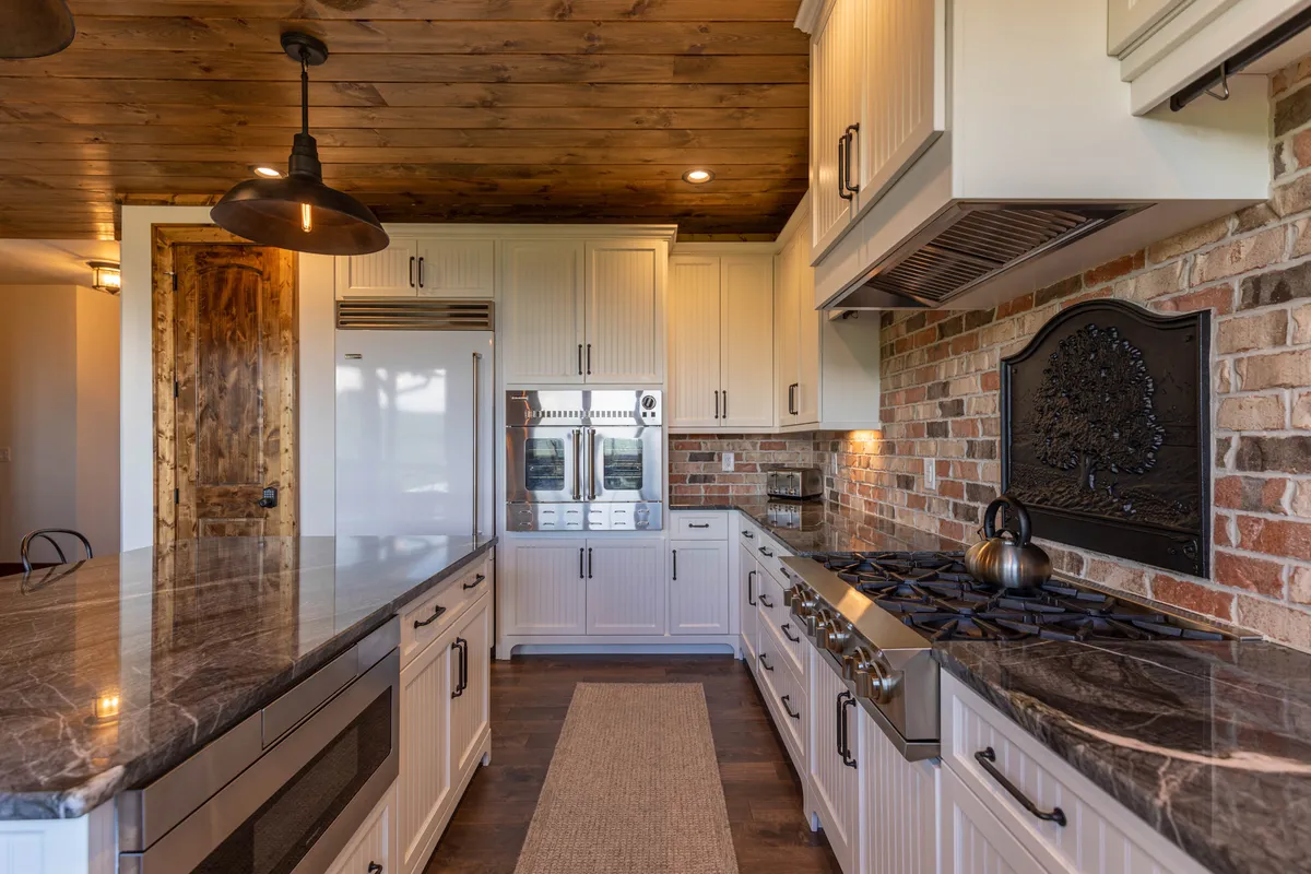 The image focuses on the gourmet kitchen, featuring gleaming granite countertops, stainless steel appliances, a brick accent wall, and a wooden ceiling. The space is clearly designed for culinary adventures and socializing.