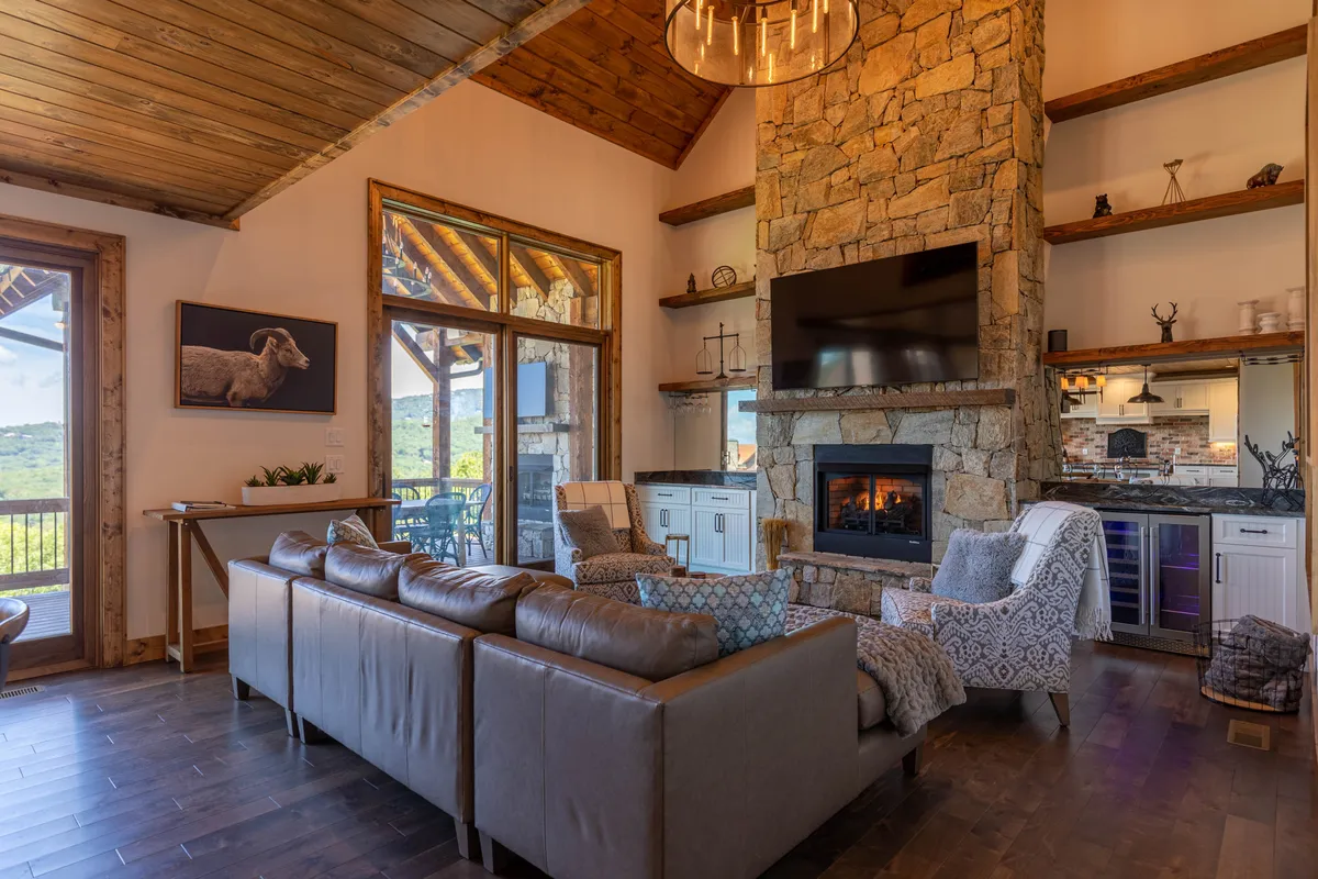 This image showcases a warm, spacious great room featuring a central stone fireplace, a comfortable sofa, and large windows that frame stunning mountain views. The TV mounted above the fireplace and the overall open design create an inviting spot for relaxation and entertainment.
