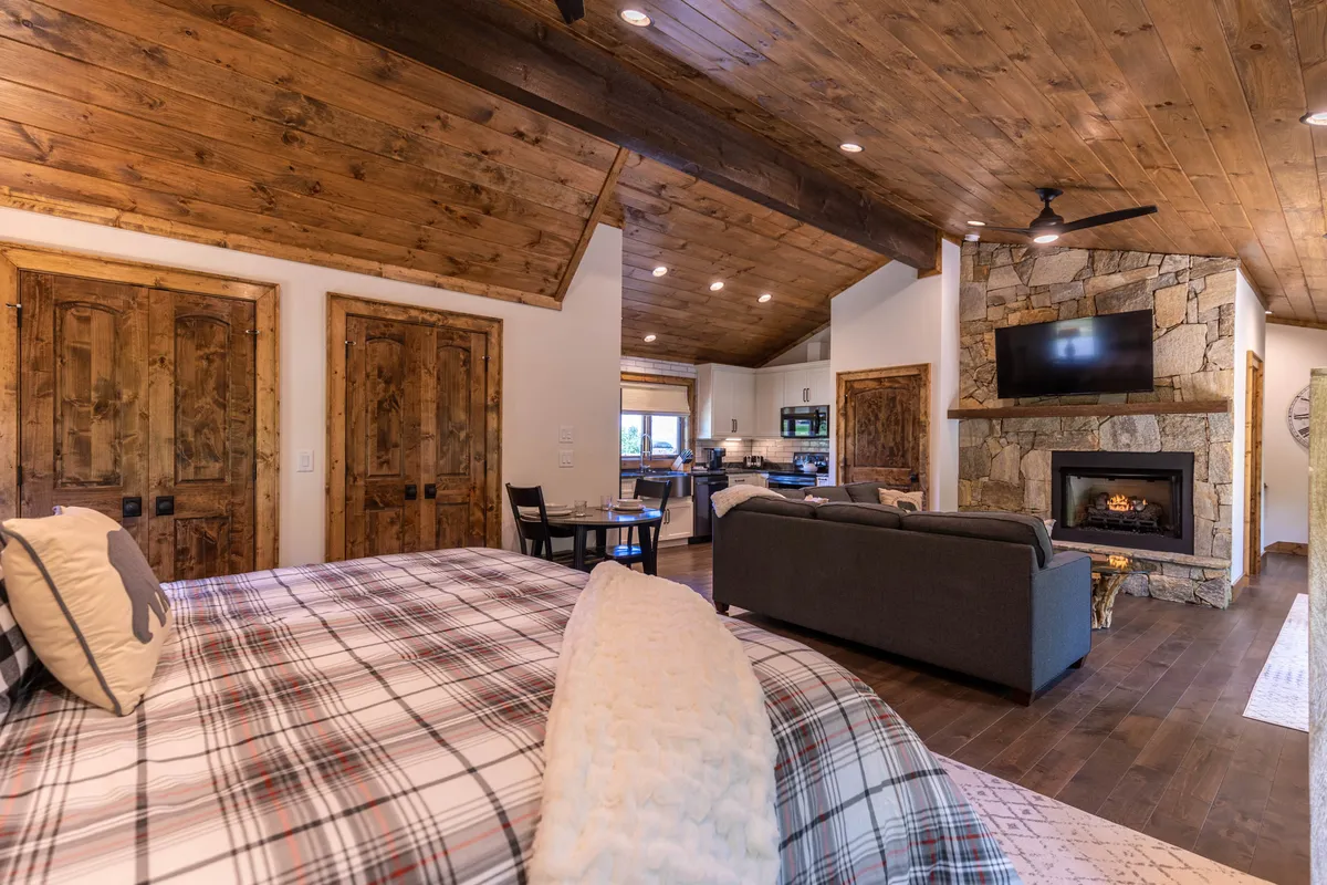 A spacious suite featuring a bed with plaid bedding, a cozy stone fireplace, wooden accents, and an open kitchen area. This area doubles as a sitting and dining space, ideal for relaxation or shared meals, and evokes the carriage house suite’s blend of privacy, comfort, and amenities.