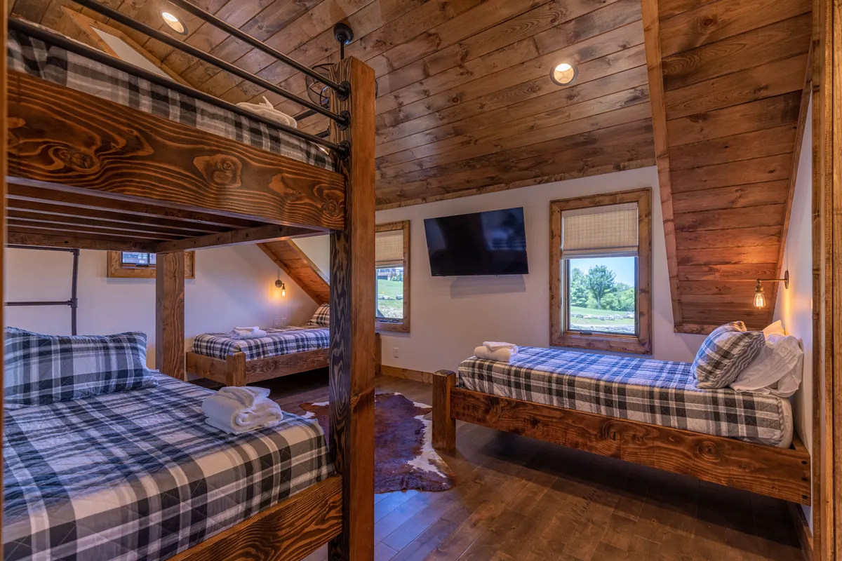 This image captures a warm, family-oriented bunk suite with both bunk beds and single beds, rustic wooden beams, a TV, and windows. The playful yet comfortable space is designed for kids or multi-generational groups, inviting storytelling and bonding.