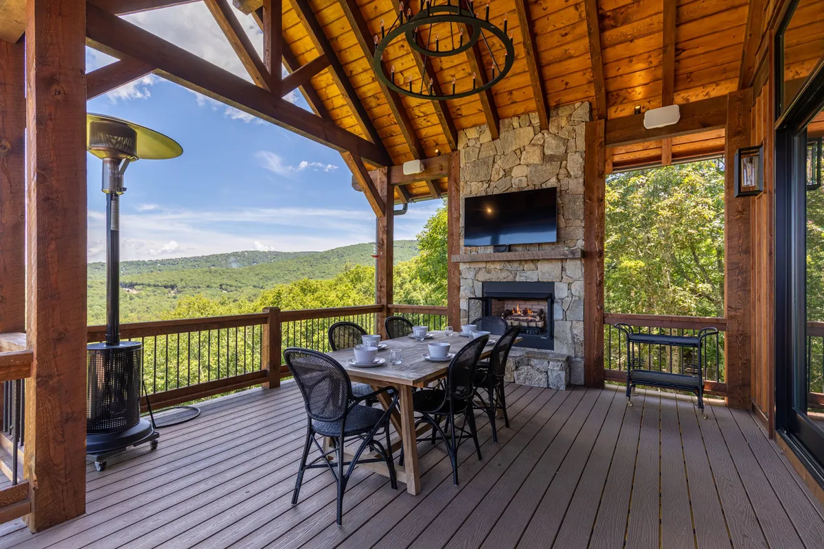 A spacious deck features a dining table set for a meal, a prominent stone fireplace, outdoor heater, decorative chandelier, and unobstructed mountain views. The warm lighting and cozy furnishings make the space ideal for gatherings in any season.