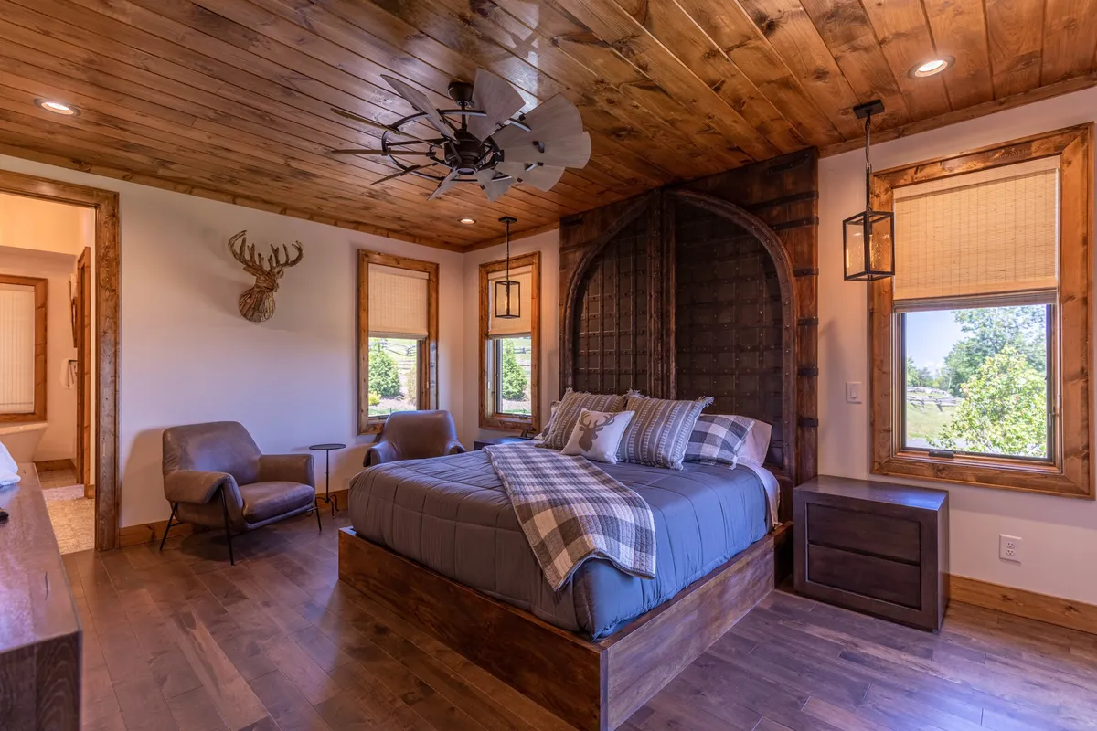 This image features a cozy bedroom with plaid bedding, a wooden wall and ceiling, large windows letting in natural light, and a comfortable armchair. The serene setup is perfect for relaxation and reflects a tranquil retreat, evoking the main-level king suite or one of the plush upstairs suites described in the section.