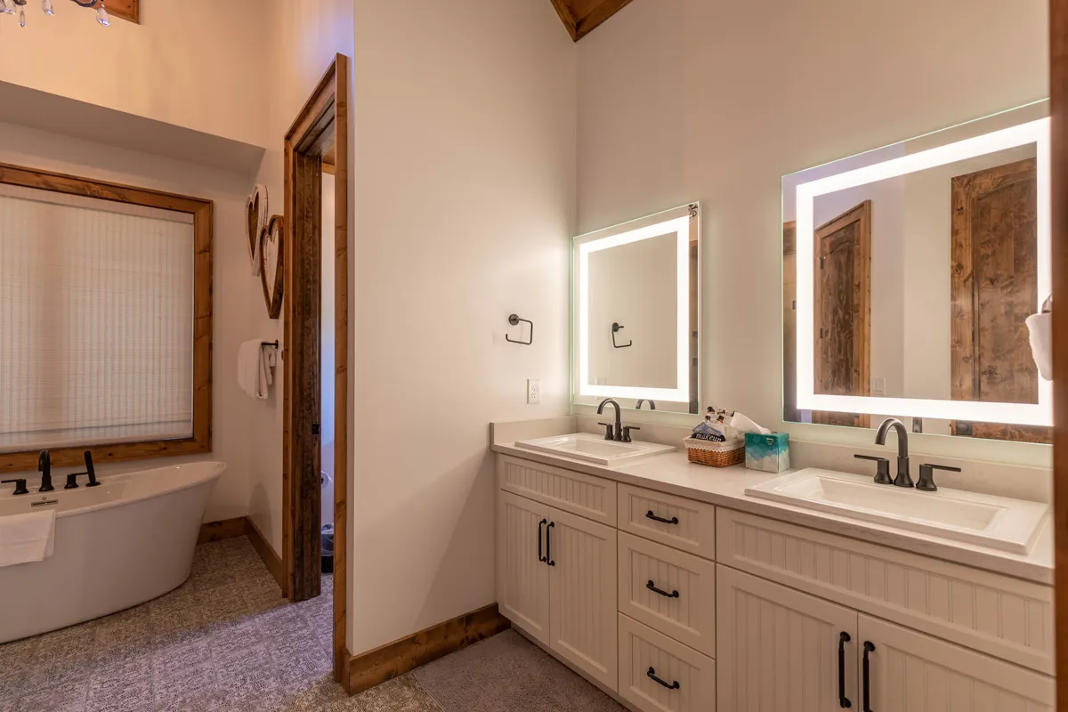 This serene bathroom features a freestanding bathtub, dual sinks with illuminated mirrors, and wooden accents. The space is calm and spa-like, ideal for unwinding or morning routines, and showcases the luxury finishes described for the main-level king suite bath and carriage house suite bath.