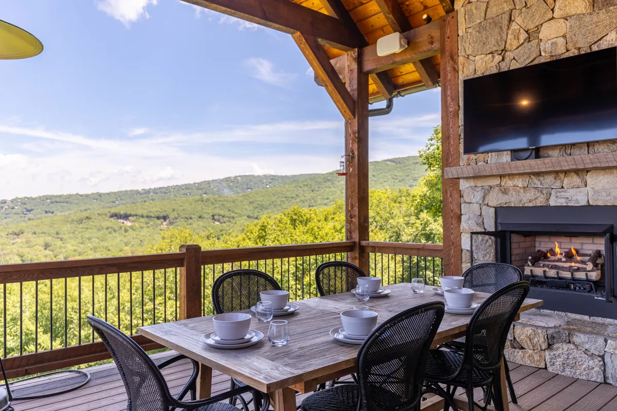 This image features an outdoor dining table set against a backdrop of stunning mountain views. Visible are comfortable outdoor seating arrangements and a cozy stone fireplace, inviting guests to relax and dine alfresco. The scene is warmly lit, evoking a sense of sociability and comfort.
