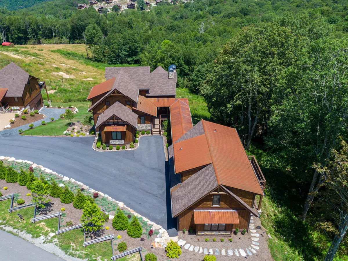 An exterior shot showcasing the main house with brown wood siding, striking red metal roofs, landscaped gardens, and a circular driveway, set in a serene mountain community. The image suggests welcoming communal spaces and the potential for holiday gatherings, family barbecues, and relaxing in a tranquil natural setting.